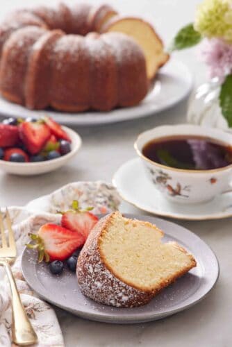 A plate with a slice of cream cheese pound cake along with some berries, dusted with powdered sugar. A drink, more berries, and the rest of the cake in the background.