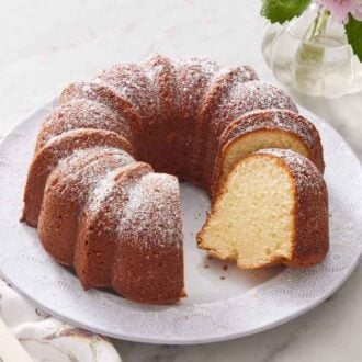 A large plate with a cream cheese pound cake with a slice cut. Powdered sugar dusted on top with a cake spatula beside the plate.