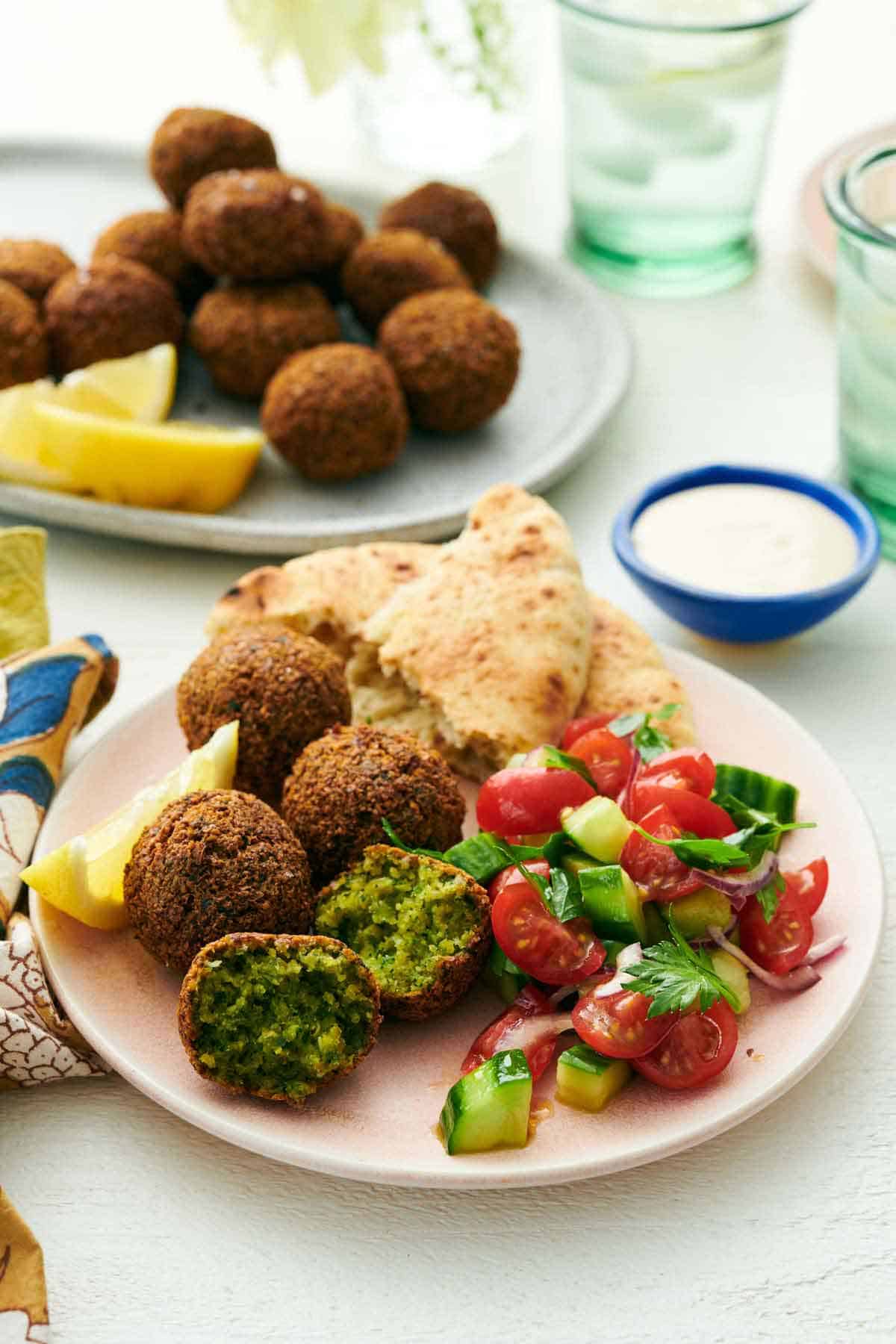 A plate with four falafel, with one cut in half. Pita bread, cucumbers, tomatoes, and a lemon wedge on the plate as well. A bowl of tahini sauce and more falafel with lemon wedges in the background.