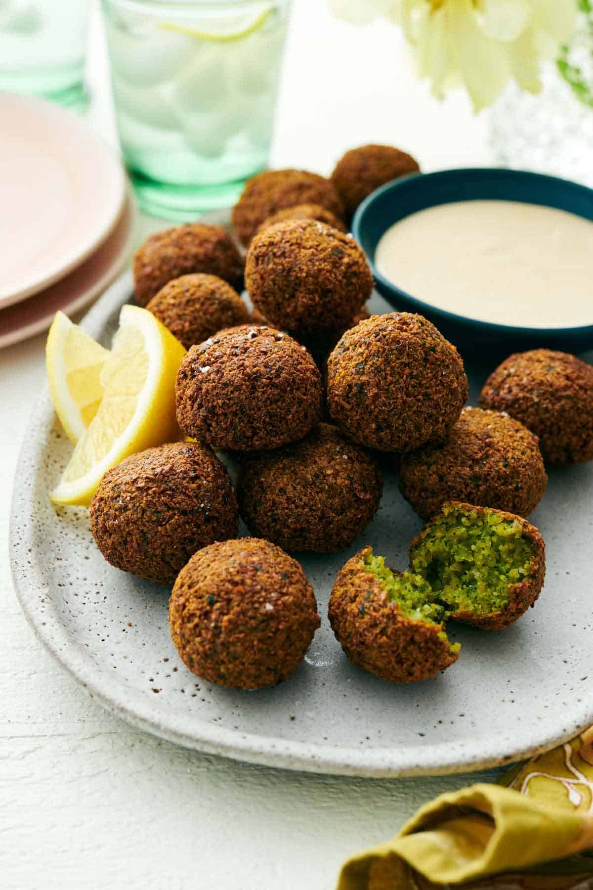 A platter with multiple falafel with lemon wedges and a bowl of tahini sauce. One falafel torn open.