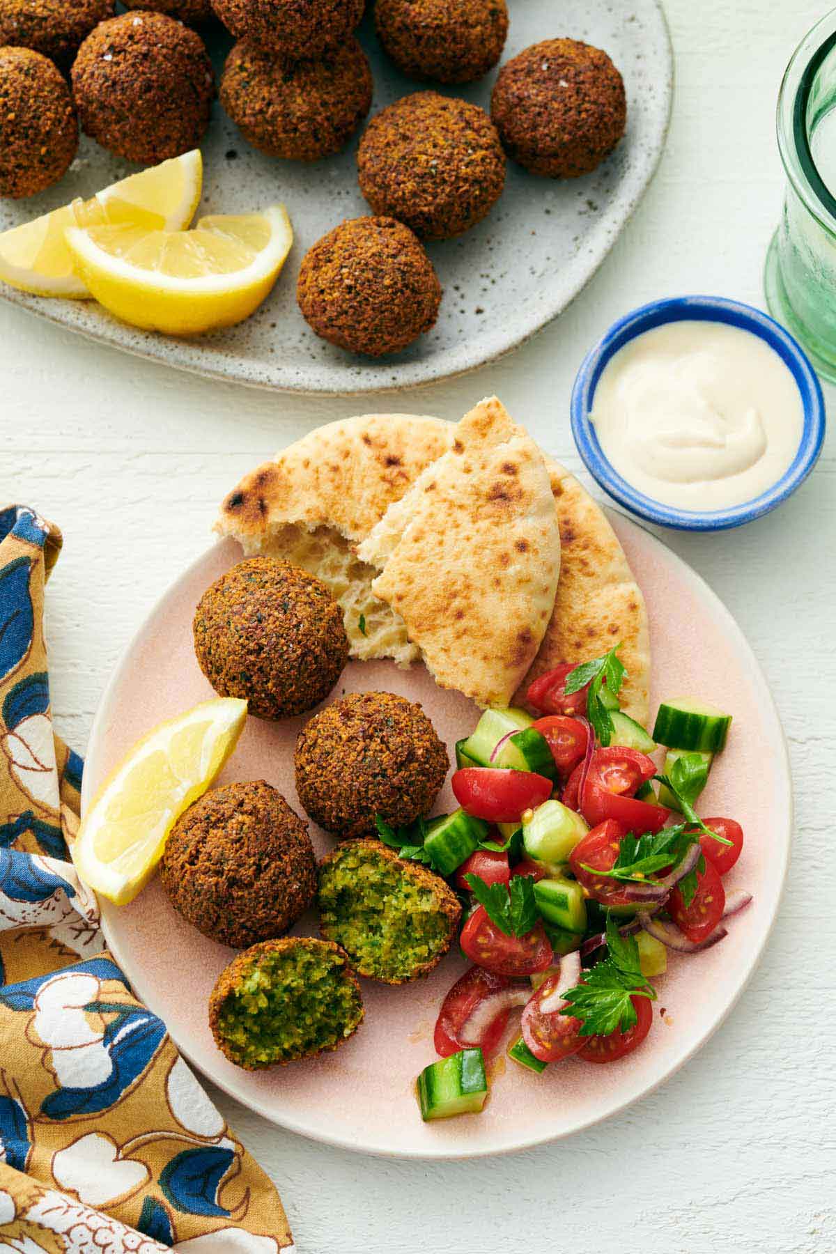 Overhead view of a plate with multiple falafel with one torn opened. Pita bread, lemon wedges, and cucumber-tomato salad. A bowl of tahini sauce and more falafel off to the side.