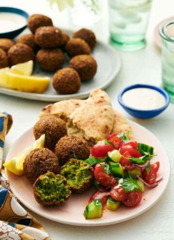 A plate with four falafel, with one cut in half, with torn pita, cucumbers, tomatoes, and a lemon wedge. A bowl of tahini sauce and more falafel with lemon wedges in the background.