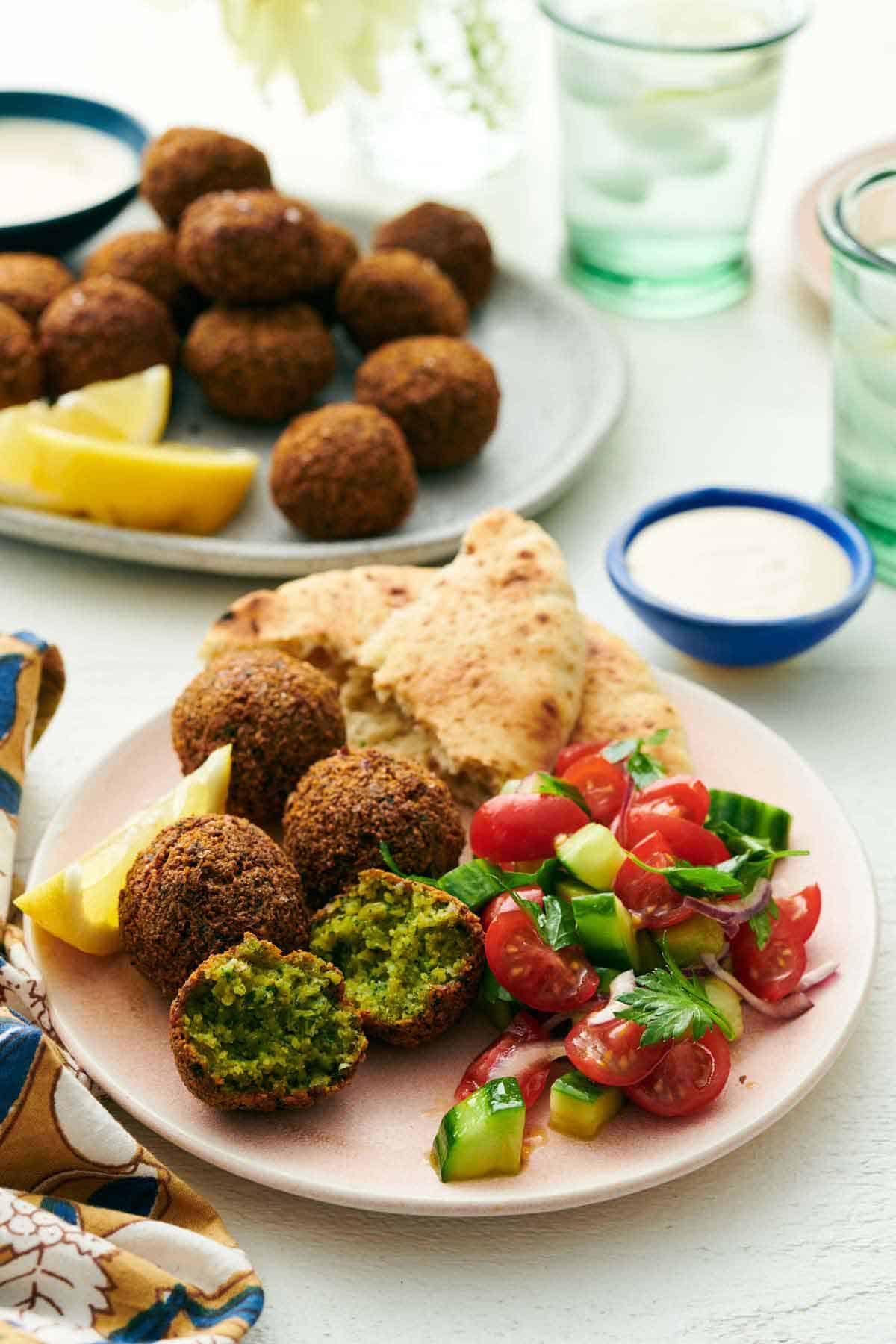 A plate with four falafel, with one cut in half, with torn pita, cucumbers, tomatoes, and a lemon wedge. A bowl of tahini sauce and more falafel with lemon wedges in the background.