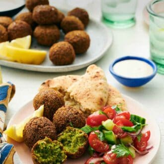 Pinterest graphic of a plate with four falafel, with one cut in half, with torn pita bread, chopped cucumbers, chopped tomatoes, and a lemon wedge. A bowl of tahini sauce and more falafel with lemon wedges in the background.