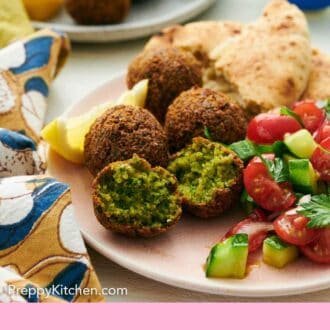 Pinterest graphic of a plate with multiple falafel with one torn opened along with torn pita bread, lemon wedges, and cucumber-tomato salad.