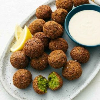 An oval platter of falafel with one torn opened. Lemon wedges and a bowl of tahini sauce on the platter.