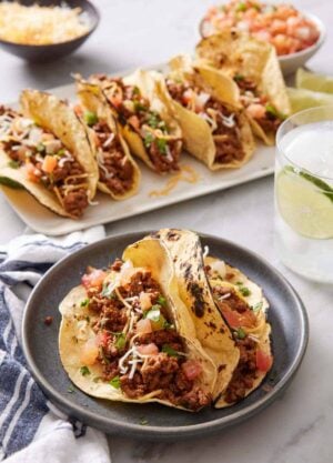 A plate with two ground beef tacos topped with cheese, pico de gallo, and cilantro. More tacos in the background along with a drink.