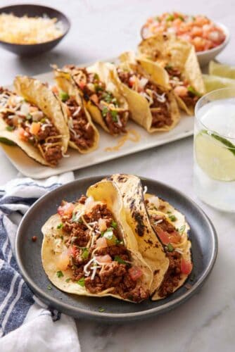 A plate with two ground beef tacos topped with cheese, pico de gallo, and cilantro. More tacos in the background along with a drink.