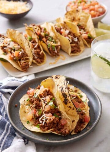 A plate with two ground beef tacos topped with cheese, pico de gallo, and cilantro. More tacos in the background along with a drink.