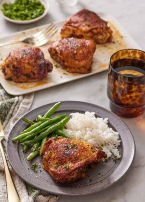 A plate with an Instant Pot chicken thigh with rice and green beans. More chicken thighs in the back on a platter along with a drink and some garnish.