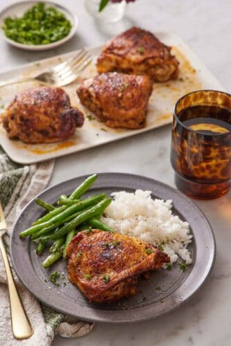 A plate with an Instant Pot chicken thigh with rice and green beans. More chicken thighs in the back on a platter along with a drink and some garnish.