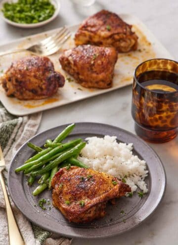 A plate with an Instant Pot chicken thigh with rice and green beans. More chicken thighs in the back on a platter along with a drink and some garnish.