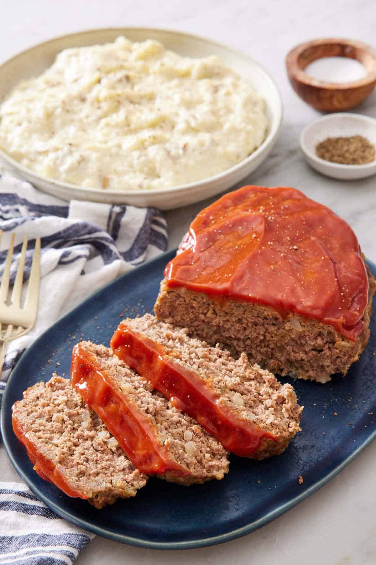 A platter with a Instant Pot meatloaf with three slices cut in front. Mashed potatoes in a bowl in the background.