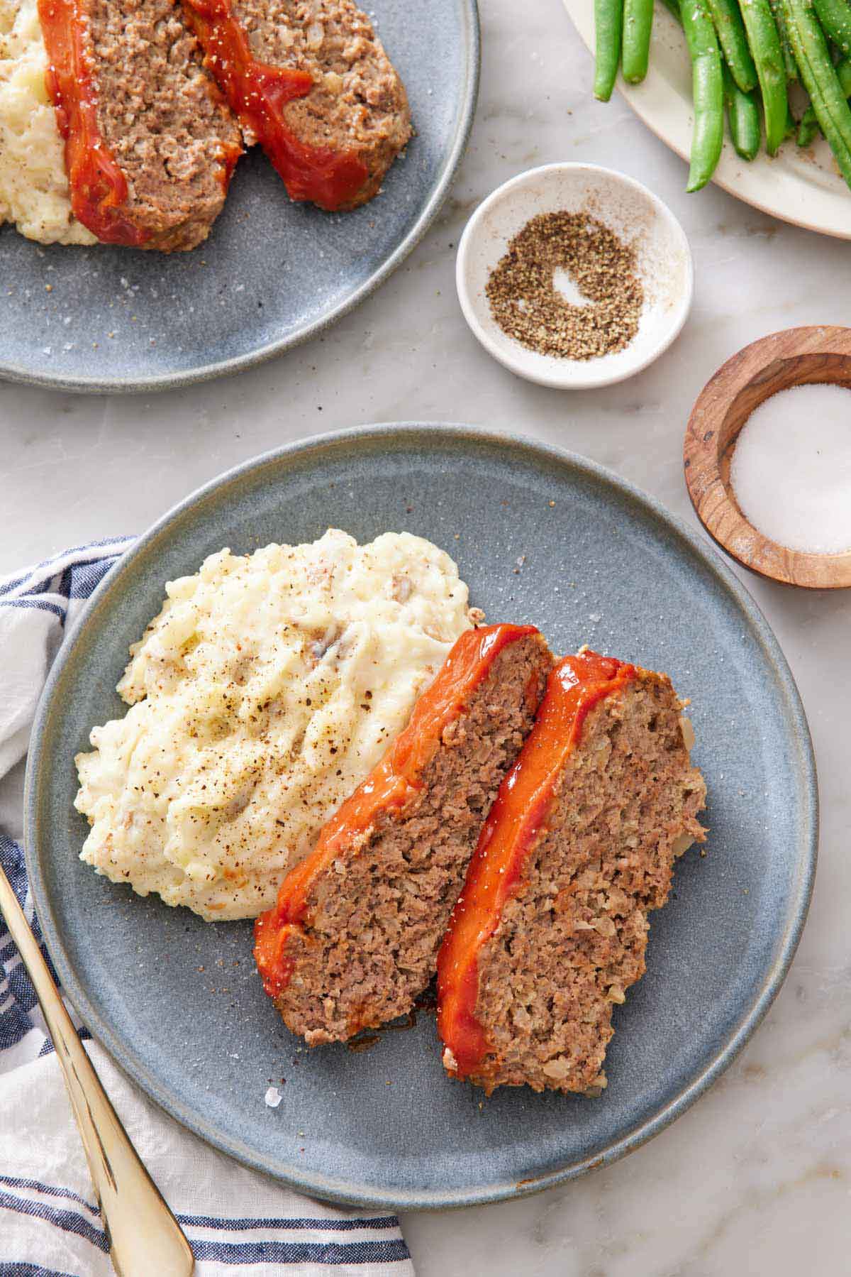 Overhead view of a plate with two slices of Instant Pot meatloaf with mashed potatoes. Salt and pepper on the side with green beans and another plate in the background.