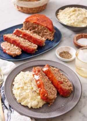 A plate with two slices of Instant Pot meatloaf with mashed potatoes. More meatloaf in the background along with a bowl of mashed potatoes, salt and pepper, and a drink.