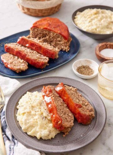 A plate with two slices of Instant Pot meatloaf with mashed potatoes. More meatloaf in the background along with a bowl of mashed potatoes, salt and pepper, and a drink.