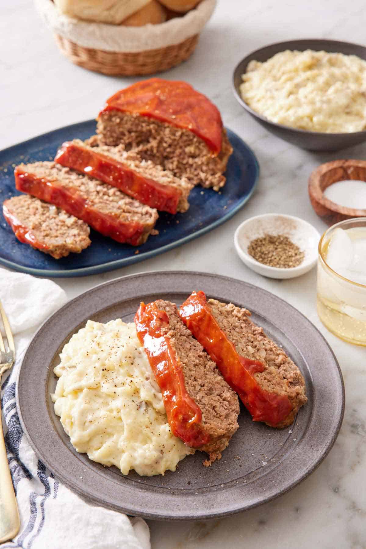 A plate with two slices of Instant Pot meatloaf with mashed potatoes. More meatloaf in the background along with a bowl of mashed potatoes, salt and pepper, and a drink.