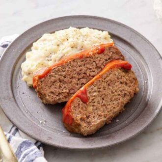 A plate with two slices of Instant Pot meatloaf and mashed potatoes. A fork and some bread on the side.