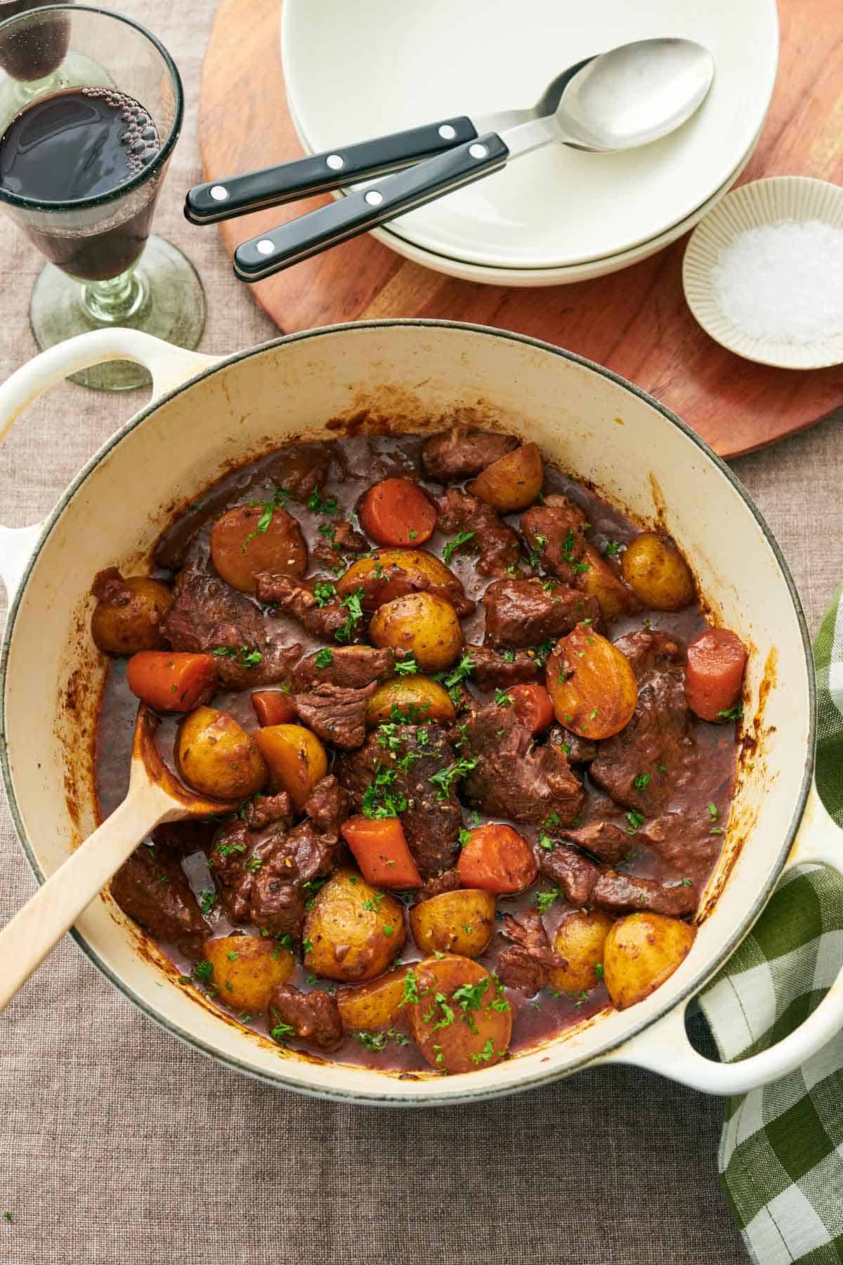 Overhead view of a pot of lamb stew with a spoon. A glass of wine, plates, spoons, and salt in the background.