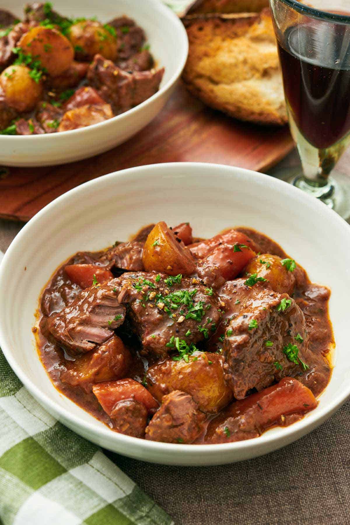 A bowl of lamb stew with chopped parsley on top. A glass of wine, bread, and another bowl of stew in the background.