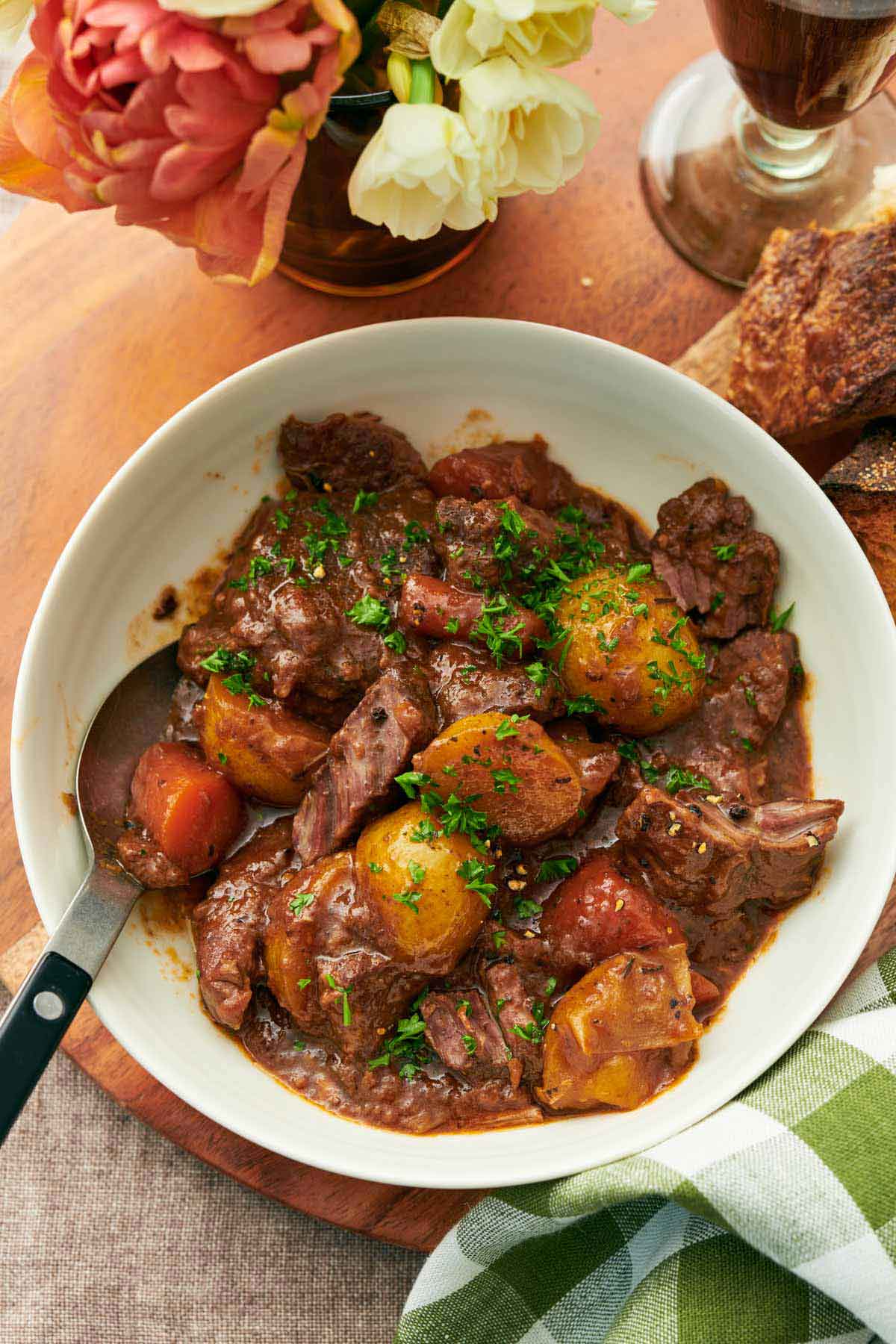 Overhead view of a bowl of lamb stew with a spoon, topped with chopped parsley. Flowers, bread, and a drink off to the side.