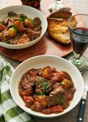 Two bowls of lamb stew with a glass of red wine and some torn bread.