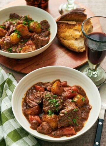 Two bowls of lamb stew with a glass of red wine and some torn bread.