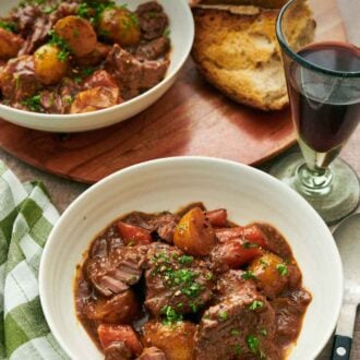 Pinterest graphic of two bowls of lamb stew with a glass of red wine and some torn bread.