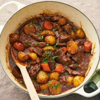 Overhead view of a white dutch oven of lamb stew with a spoon. A green checkered linen napkin on the side.