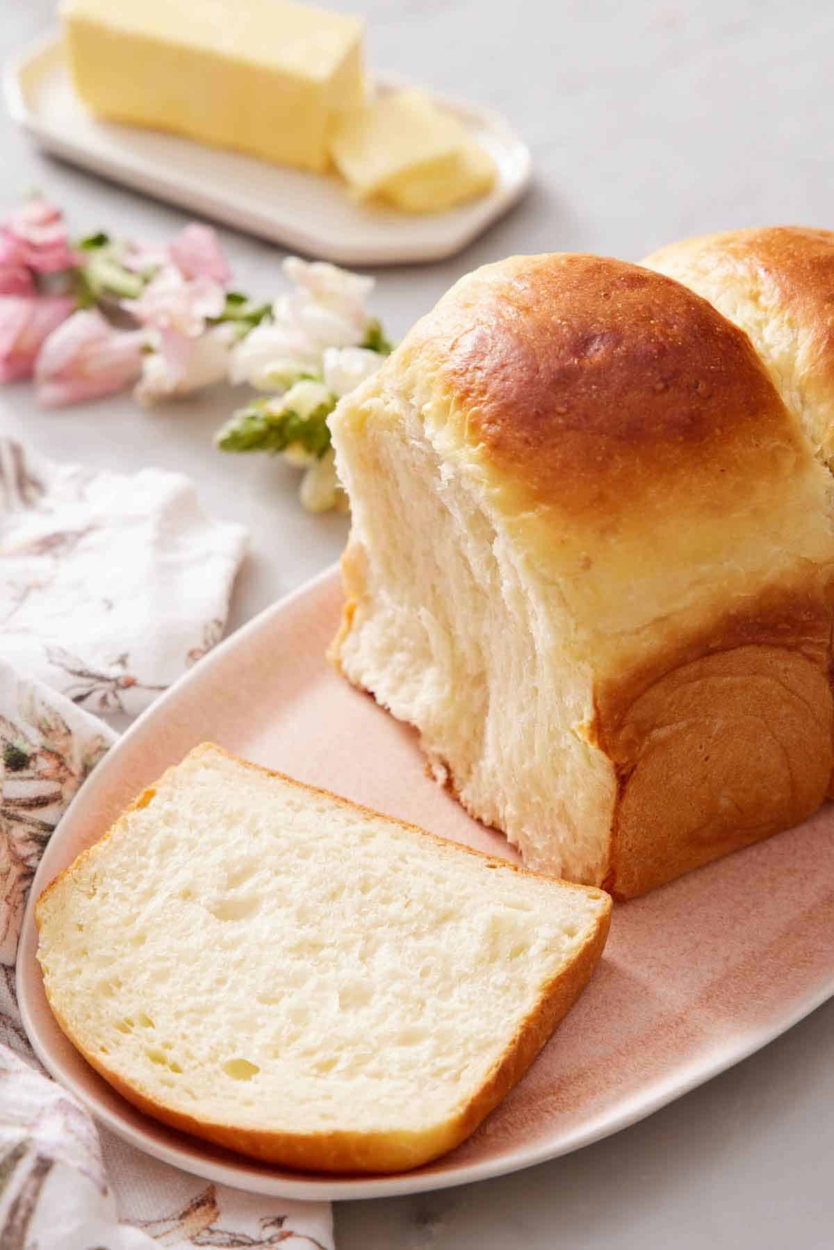 A platter with a loaf of milk bread with a slice cut in front. A cut stick of butter in the background.