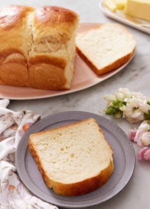 A plate with a slice of milk bread. The rest of the loaf in the back on a platter with one slice cut.