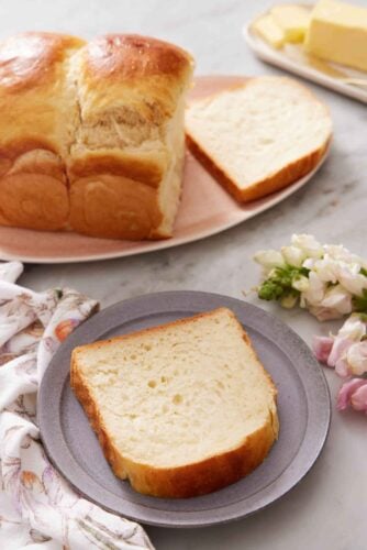 A plate with a slice of milk bread. The rest of the loaf in the back on a platter with one slice cut.