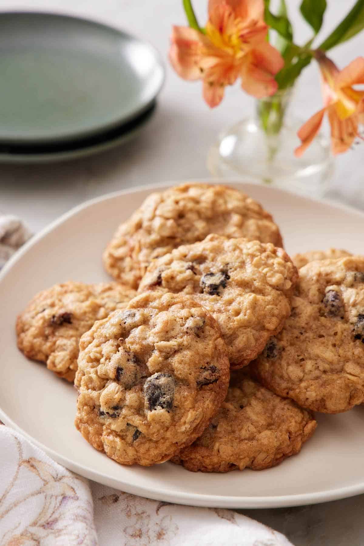 A platter with a pile of oatmeal cookies. Flowers in the background with a stack of two plates.