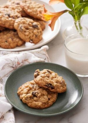 A green plate with two oatmeal cookies with a glass of milk, flowers, and platter of more cookies in the background.