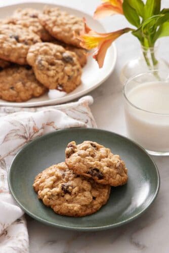 A green plate with two oatmeal cookies with a glass of milk, flowers, and platter of more cookies in the background.