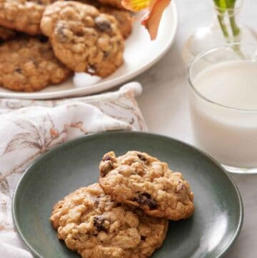 A green plate with two oatmeal cookies with a glass of milk, flowers, and platter of more cookies in the background.