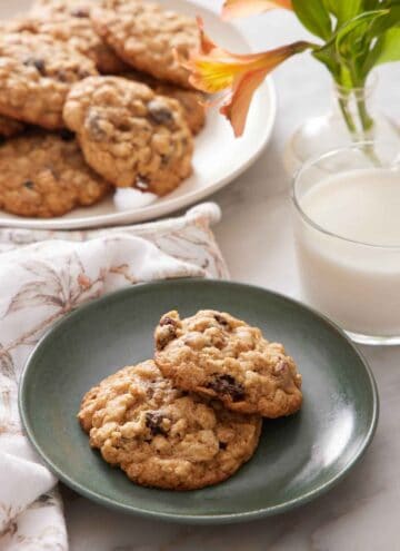 A green plate with two oatmeal cookies with a glass of milk, flowers, and platter of more cookies in the background.