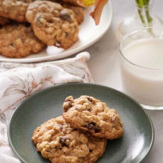 Pinterest graphic of a plate with two oatmeal cookies with a glass of milk, flowers, and platter of more cookies in the background.