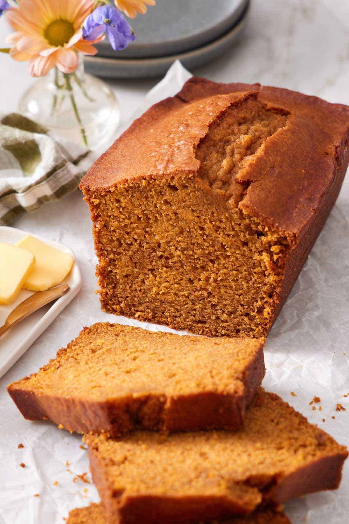 A loaf of pumpkin bread with three slices cut in front of it. Butter, flowers, and plates in the back.