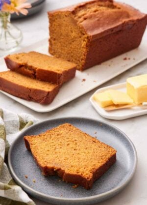 A plate with a slice of pumpkin bread. A loaf in the background with two slices cut in from to fit. Butter on the side.