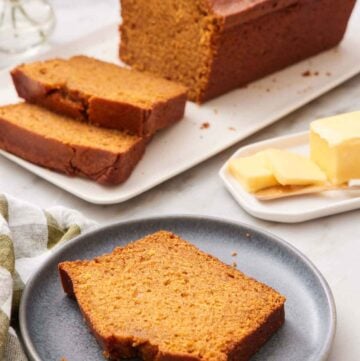 A plate with a slice of pumpkin bread. A loaf in the background with two slices cut in from to fit. Butter on the side.