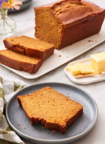 A plate with a slice of pumpkin bread. A loaf in the background with two slices cut in from to fit. Butter on the side.