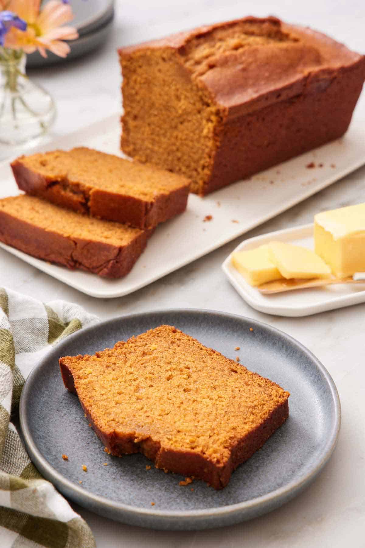 A plate with a slice of pumpkin bread. A loaf in the background with two slices cut in from to fit. Butter on the side.