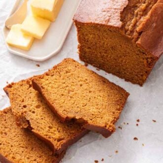 Three slices of pumpkin bread in front of the loaf with some butter on the side, all on top of parchment paper.