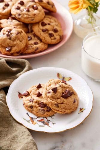 A plate with two pumpkin chocolate chip cookies with a glass of milk and platter of more cookies in the background.