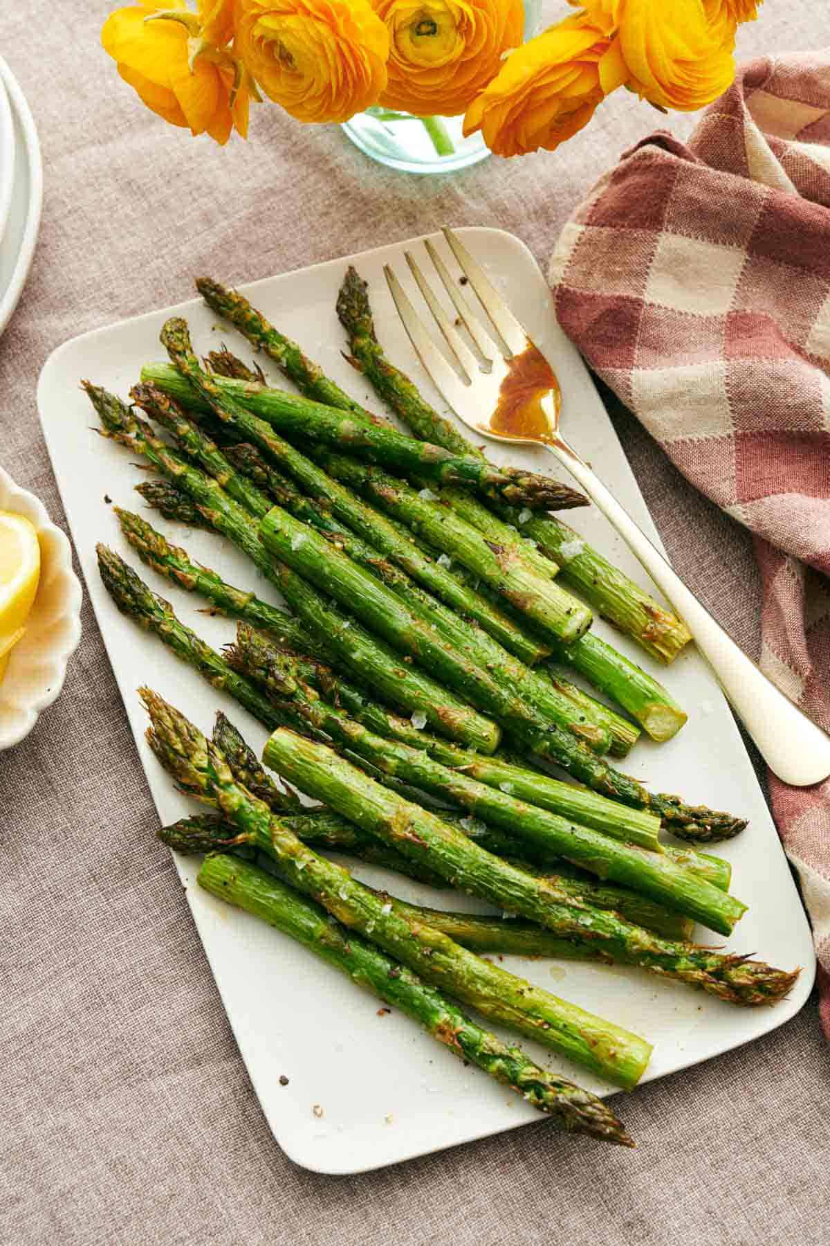 A platter of roasted asparagus with a serving fork. Flowers and a linen napkin on the side.