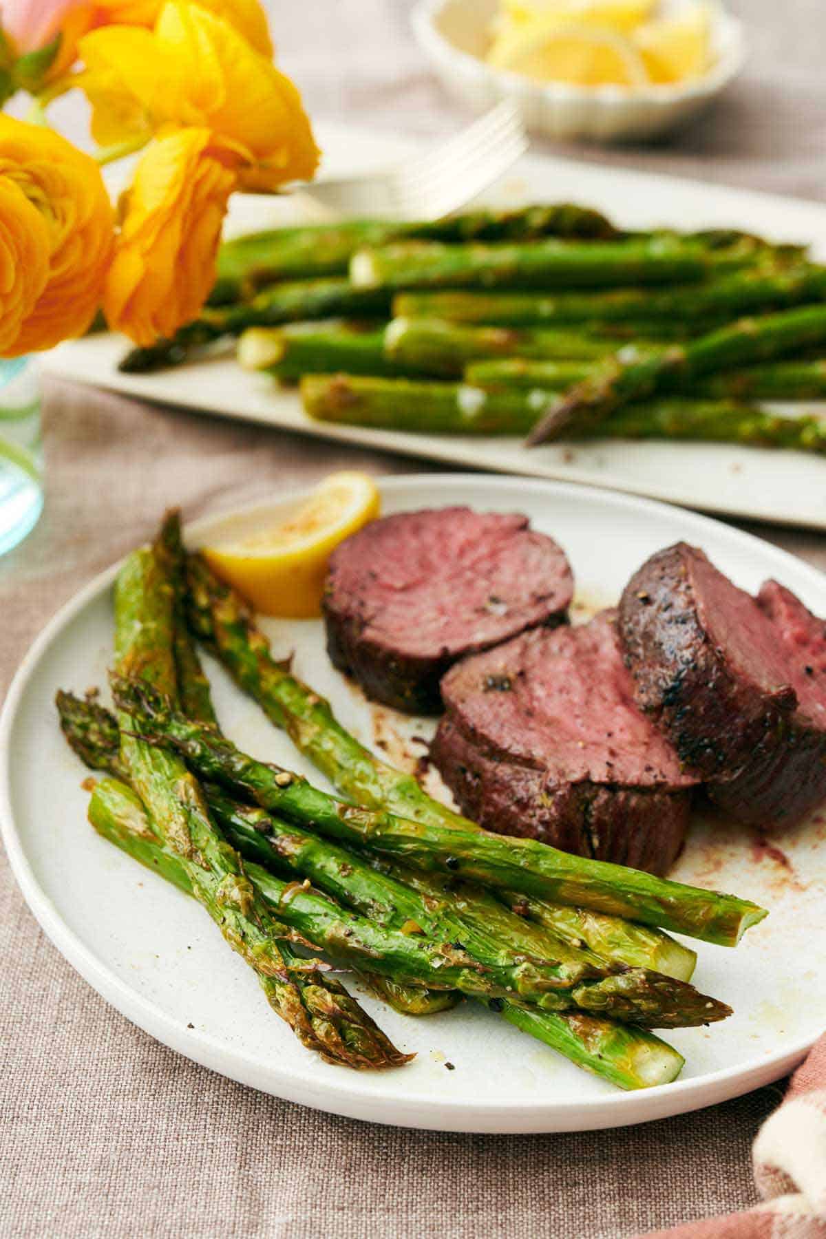 A plate with roasted asparagus, steak, and a lemon wedge. More asparagus in the background on a platter.