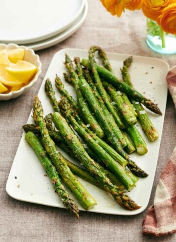 A platter of oven roasted asparagus. Lemon wedges, stack of plates, and flowers in the background.