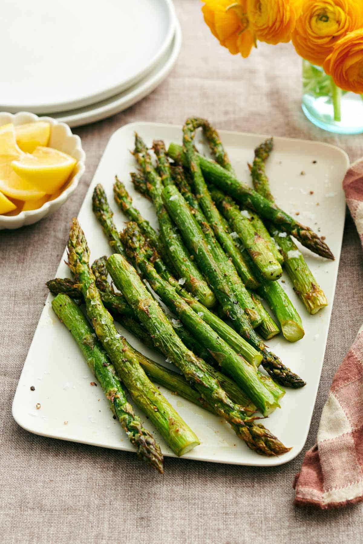 A platter of oven roasted asparagus. Lemon wedges, stack of plates, and flowers in the background.