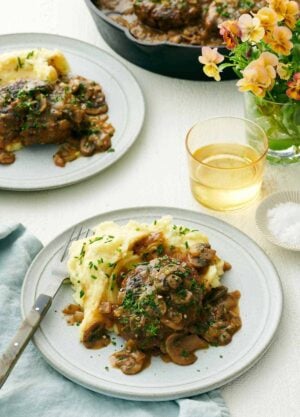 A plate with salisbury steak and mashed potatoes. A second plated serving in the back along with a drink, vase of flower, and skillet.